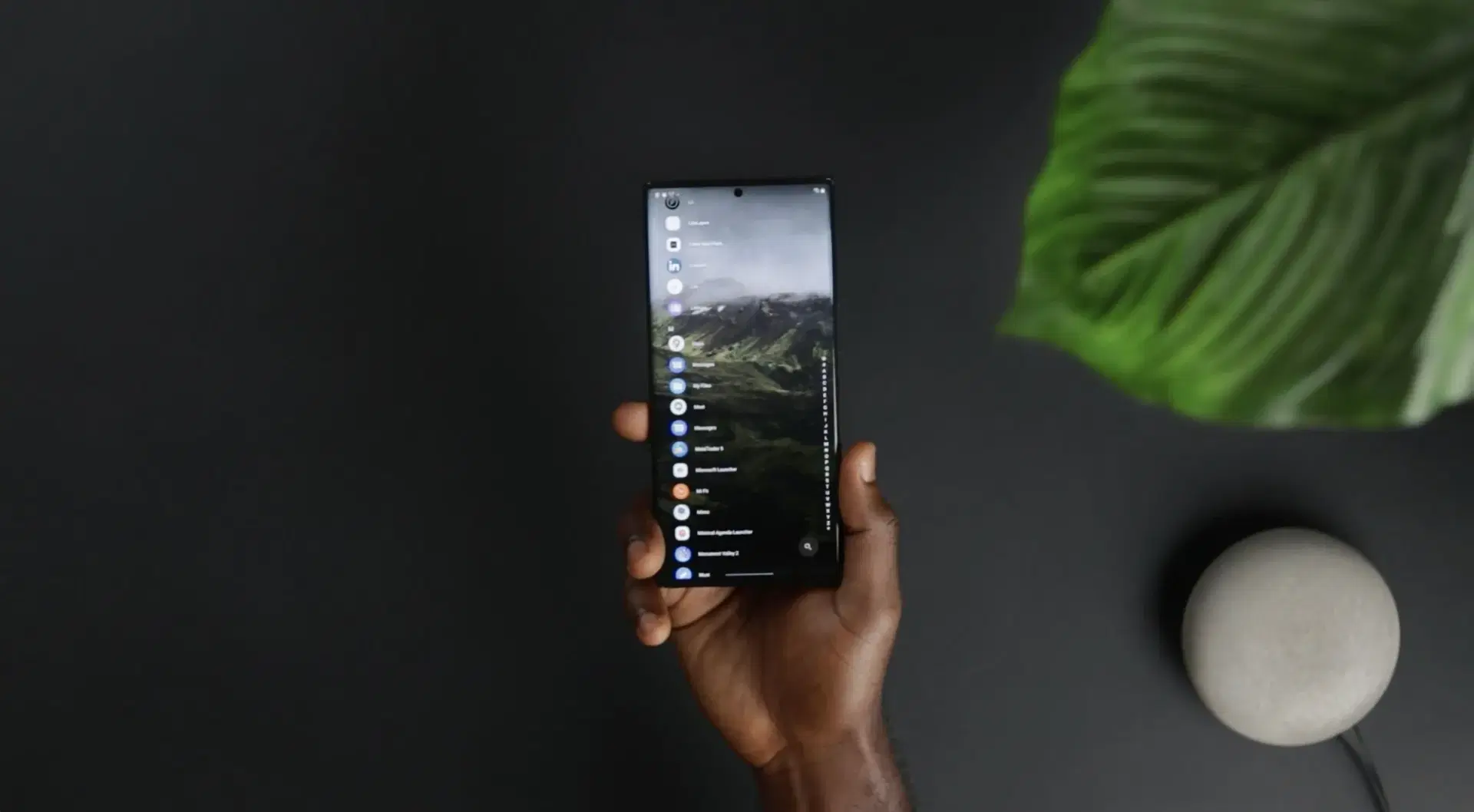 A hand holding a phone with an app list open, on a dark table