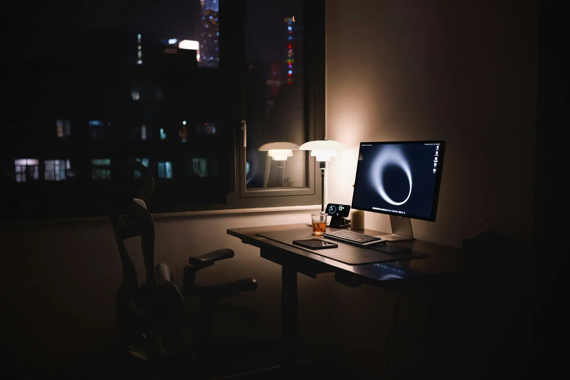 A desk with a monitor and a lamp by a window at night, city lights outside